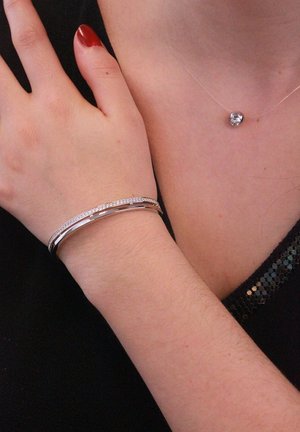 Braccialetto d'argento con un design curvo, impreziosito da gemme trasparenti lungo il bordo superiore. Indossato su un polso accanto a un semplice collier d'argento con un pendente rotondo.