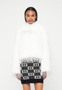 Bela umetna krzna jakna z visokim ovratnikom in frufru robovi, v kombinaciji z črnimi krili, ki imajo bel geometrijski vzorec.