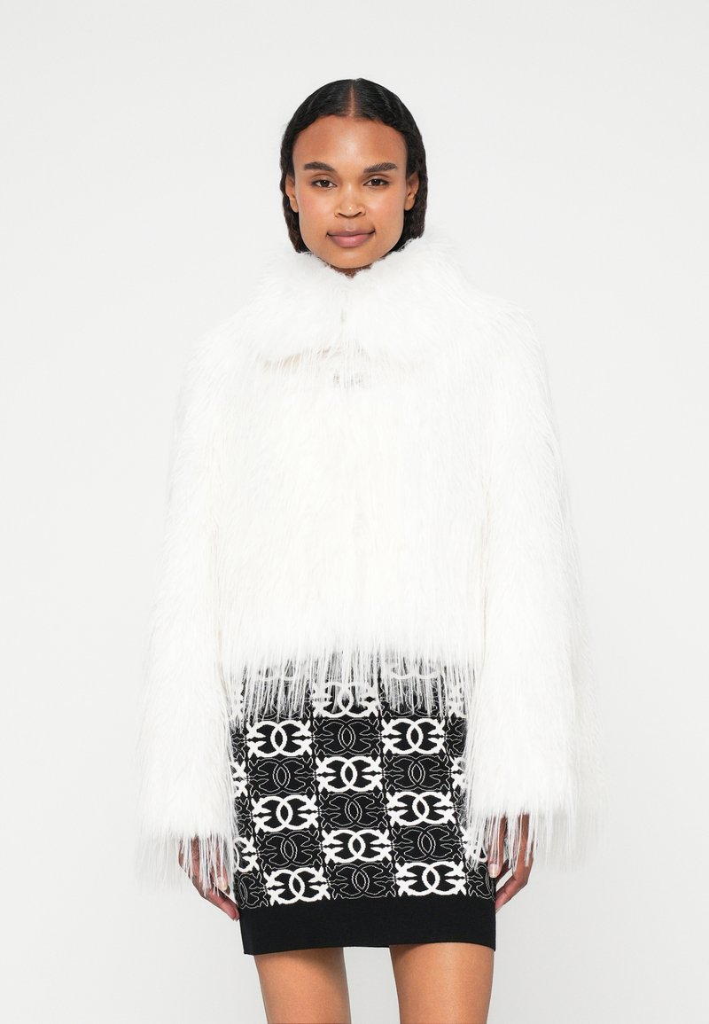 Bela umetna krzna jakna z visokim ovratnikom in frufru robovi, v kombinaciji z črnimi krili, ki imajo bel geometrijski vzorec.