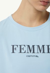 Hellblaues Baumwoll-T-Shirt mit Rundhalsausschnitt und kurzen Ärmeln. Enthält "FEMME" in fettem, schwarzem Text, mit dem kleineren "comma," darunter.