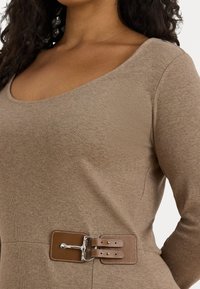 Nærbilde av en person som har på seg en beige strikket topp med bred utringning og et brunt lærbelte med dobbelt hodefeste og sølvspenne i livet.