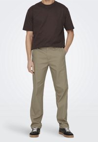 Brązowy T-shirt z krótkim rękawem, zestawiony z jasnobeżowymi spodniami oraz czarnymi trampkami z białymi sznurowadłami i brązowymi podeszwami. Casual, luźny fason.