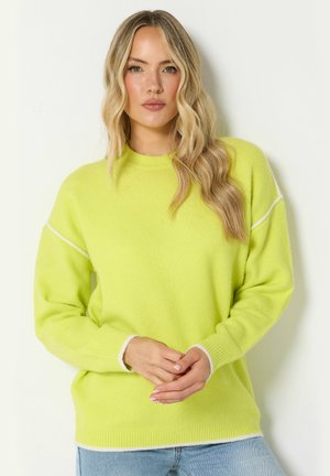 Femme aux longs cheveux blonds ondulés portant un pull jaune vif avec des bordures blanches et un jean bleu clair, debout devant un fond blanc.