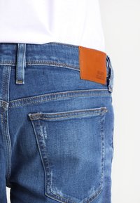 Jeans i ljusblå denim med en brun läderlappar vid midjan präglad med "RIVER ISLAND." Har subtil slitage och flera fickor.