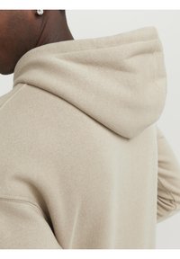 Sudadera beige con una textura suave, que presenta una capucha con cordón y puños acanalados. La tela parece lisa y puede tener un ligero brillo.