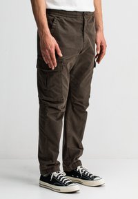 Bruine cargobroek met een elastische tailleband, voorzien van meerdere zijzakken, gestyled met zwarte sneakers en een wit T-shirt.