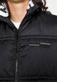 Zbliżenie na osobę w czarnej kurtce puchowej z logo "THE COUTURE CLUB" na piersi i zamkiem błyskawicznym z przodu, częściowo rozpiętym.