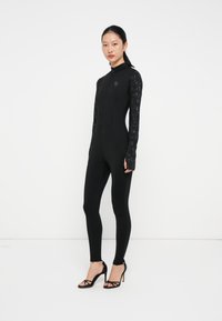 Svart heldekkende bodysuit med lange blondermerker, med høy krage og glidelås. Utsmykket med en liten logo på brystet.
