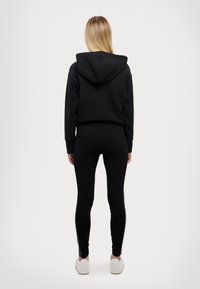Črna prešita kapuca s kapuco, v kombinaciji s prilegajočimi črnimi leggings. Obleko dopolnjujejo bele teniske.