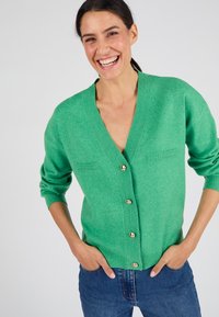 Groene wollen cardigan met een diepe V-hals, knoopsluiting en twee voorzakken. Voorzien van goudkleurige knopen en geribbelde manchetten.