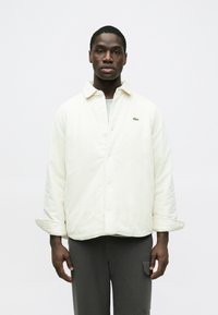 Krémfehér kordbársony ingdzseki laza szabással, gombos lezárással és egy kis zöld logóval. Sötétszürke nadrággal párosítva.