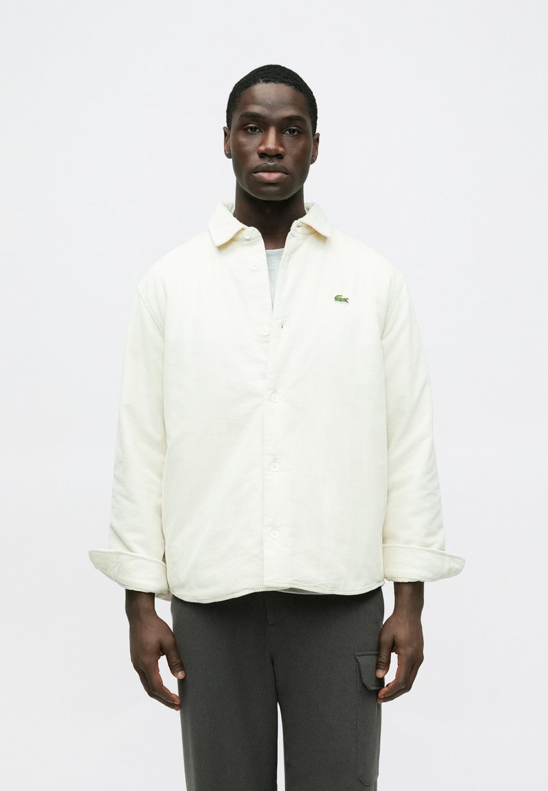 Krémfehér kordbársony ingdzseki laza szabással, gombos lezárással és egy kis zöld logóval. Sötétszürke nadrággal párosítva.