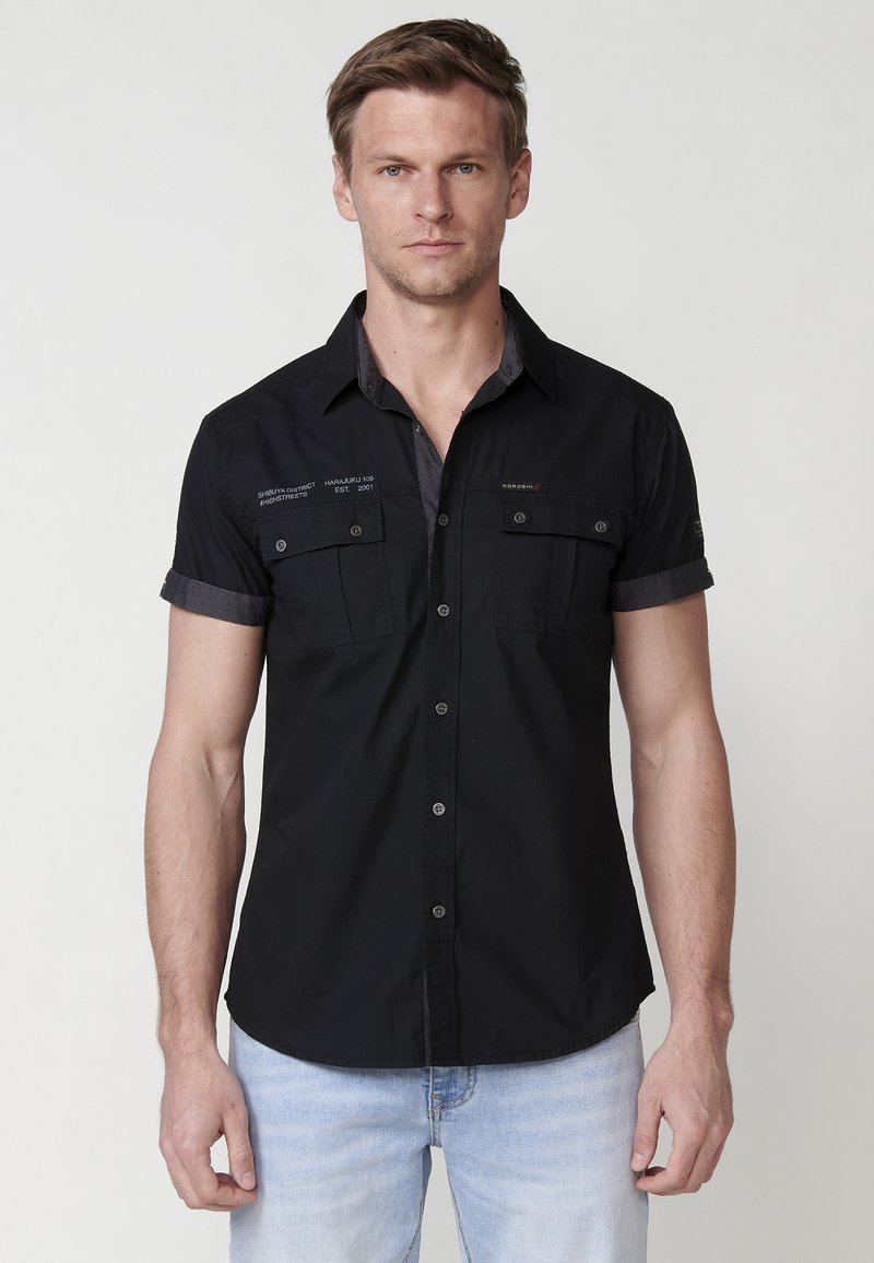 Camisa negra de mangas cortas con dos bolsillos en el pecho, cuello oscuro en contraste, cierre de botones al frente y sutil detalle de logo en la tela.