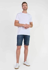 Camiseta blanca de algodón, pantalones cortos de mezclilla azul con dobladillos enrollados y zapatillas blancas con suelas beige. El modelo se encuentra de pie contra un fondo liso.