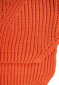 Neuletta oranssia kangasta, jossa on tekstuurillinen kuviointi, joka esittää pystysuoria ja vinoviivoja, ja jossa näkyy tiheä kudonta sekä ribattu yksityiskohta.