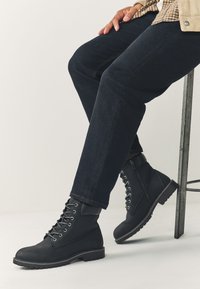 Čierne členkové topánky z textúrovaného materiálu s kovovými očkami, šnúrkami, bočným zipsom a robustnou gumovou podrážkou. Nosené s tmavými džínsami.