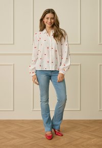 Jonge vrouw lacht, draagt een witte blouse met rode hart- en rozenpatronen, lichtblauwe spijkerbroek, rode schoenen, staand op een houten vloer tegen een betegelde muur.