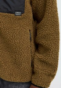 Brown fleece jacket with black zippered pocket and black patch label on the chest, worn over dark pants.