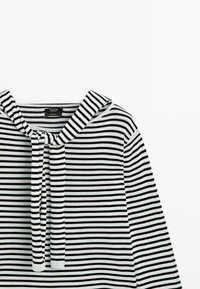 Sort og hvid, vandret striped langærmet skjorte med bundet stofdetalje omkring halsudskæringen, vist på en hvid baggrund.