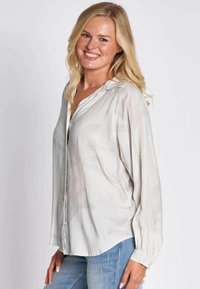 Hellgraue Bluse mit Kapuze, vorne mit Knöpfen geschlossen und langen Ärmeln. Dezente rosa Muster und strukturierte Stoffdetails sind vorhanden.