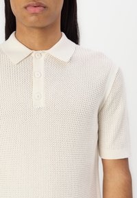 Strikket polo skjorte i en lys kremfarge med en teksturert, åpen vev. Har krage i klassisk stil og knappestolpe, korte ermer med ribbestrikkede mansjetter.