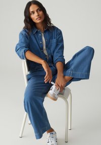 Tenue en denim comprenant une chemise bleue à boutons avec une poche poitrine et un pantalon large. Baskets blanches avec des accents gris et des semelles beige clair.