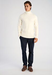 Krämfärgad polotröja i mjukt stickat material, kombinerad med mörkblå byxor och bruna läderskor med snörning och texturerad sula.