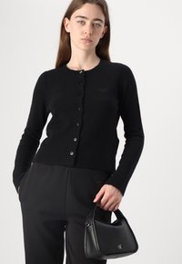 Cardigan nero in lana con maniche lunghe, orlo a coste e chiusura con bottone. Abbinato a pantaloni neri e una piccola borsa nera.