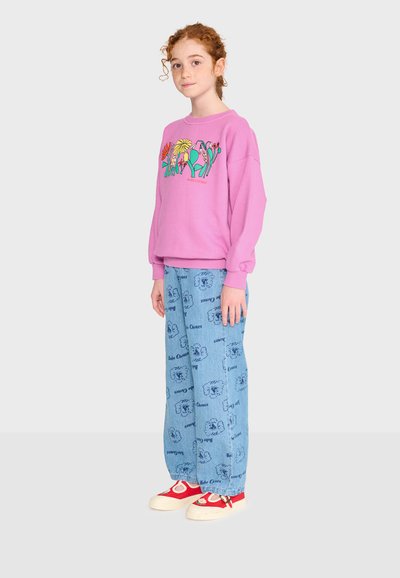 Jeune personne aux cheveux roux bouclés portant un sweat-shirt rose avec un motif floral coloré, un jean bleu à motifs et des baskets rouges.