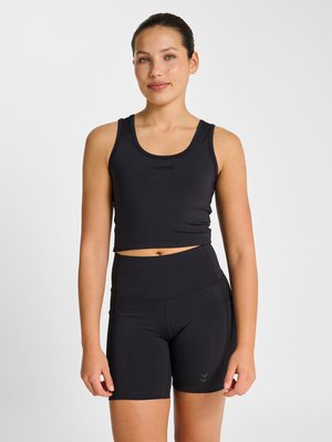 Ung kvinde iført sort ærmeløs crop top og højtaljede biker-shorts, stående mod en ensfarvet hvid baggrund.