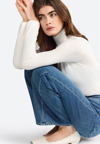 White knit turtleneck top with specks, light blue wide-leg jeans, and cream-colored block-heel shoes. Model seated with knees drawn up.