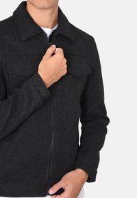 Giacca in pile nera con zip frontale, colletto a risvolto, due tasche sul petto e una finitura testurizzata. Il modello tiene la zip con una mano.