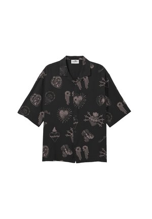 Giovane con capelli bagnati pettinati all'indietro, indossa una maglietta nera a maniche corte con disegni rosa, pantaloni bianchi e scarpe nere, in piedi in posizione neutra.