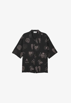 Giovane con capelli bagnati pettinati all'indietro, indossa una maglietta nera a maniche corte con disegni rosa, pantaloni bianchi e scarpe nere, in piedi in posizione neutra.