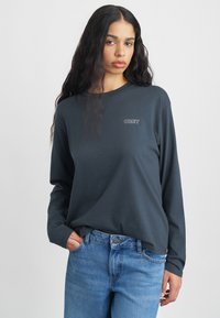 Ciemnoszara bluzka z długim rękawem i okrągłym dekoltem, z małym białym logo "OBEY" po lewej stronie klatki piersiowej. Połączona z jasnoniebieskimi dżinsami.