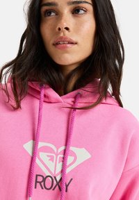Rosa hoodie med dragsko, med en vit hjärta- och diamantgrafik samt "ROXY"-text. Mjukt material, avslappnad design, medelhög tjocklek.