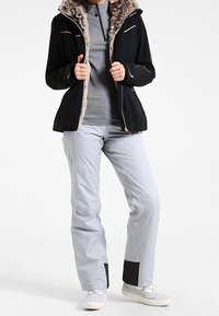 Schwarze Skijacke mit Kunstpelzkapuze, Reißverschlussakzenten und einem enganliegenden grauen Oberteil, kombiniert mit hellgrauen Skihosen mit schwarzen Details und weißen Schuhen.