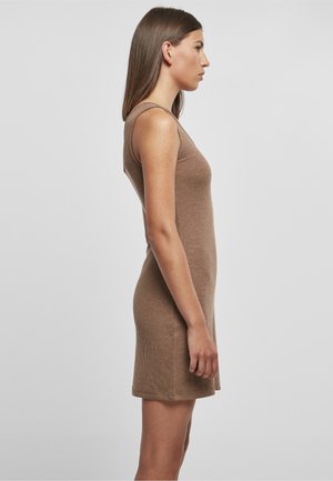 Junge Frau mit langen braunen Haaren, die ein ärmelloses, figurbetontes, knielanges beiges Strickkleid trägt und im Profil vor einem einfarbigen grauen Hintergrund steht.