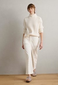 Witte geribbelde coltrui gecombineerd met crèmekleurige wijde broek. Model draagt grijze sandalen. Gladde texturen, neutraal kleurenpalet.