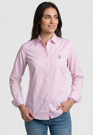Mujer que lleva una camisa rosa claro abotonada con mangas largas y un pequeño logo azul bordado en el pecho, combinada con vaqueros azules.