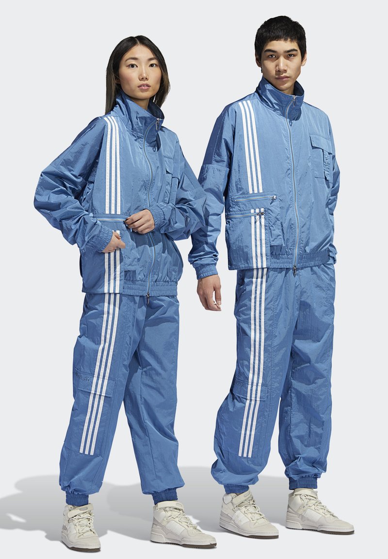 Blauwe trainingspakken met witte strepen, voorzien van ritszakken, elastische boorden en een gladde, lichte textuur. Draag door twee modellen.