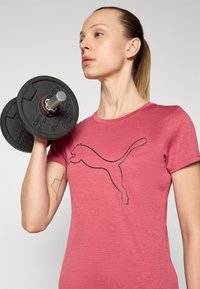 Rosa kortermet t-skjorte med en svart Puma-logo, båret av en person som holder en svart manual. Glatt stofftekstur.