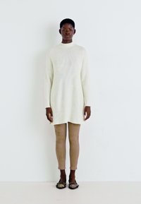 Robe en tricot blanche à manches longues, col montant et texture côtelée ; associée à des leggings marron clair et des ballerines noires à motifs.
