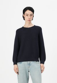 Ναυτικό πλεκτό πουλόβερ με στρογγυλή λαιμόκοψη, μακριά μανίκια και ριχτό τελείωμα, συνδυασμένο με ανοιχτό μπλε τζιν σε χαλαρή εφαρμογή.