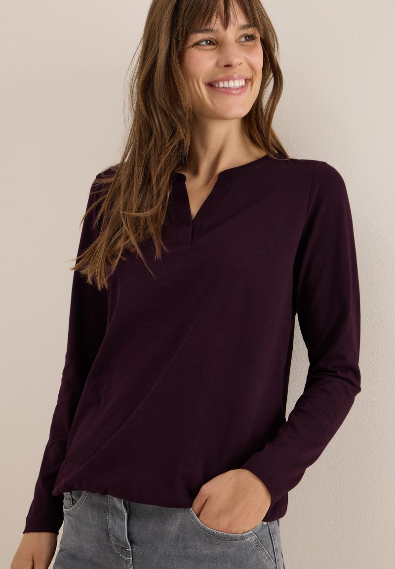 Langarmshirt in tiefem Lila, mit V-Ausschnitt und weicher Textur. Kombiniert mit hellgrauem Denim. Schlichtes und lässiges Design.