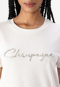 Bílý bavlněný tričko s texturovaným nápisem "Champagne" v metalických akcentacích přes hruď. Klasický styl s kulatým výstřihem.