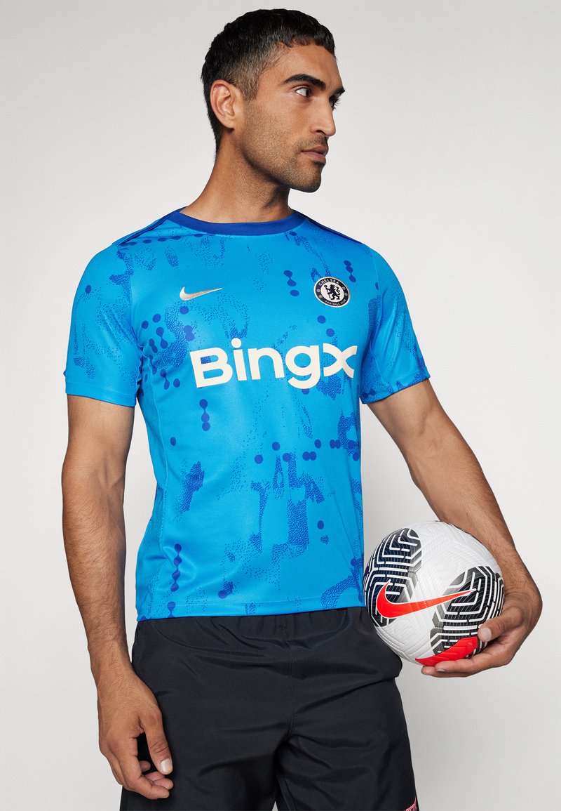 Niebieska koszulka piłkarska z abstrakcyjnymi wzorami i logo Nike; zawiera tekst "Bingx", herb Chelsea oraz okrągłą piłkę do footballu w czarno-białych kolorach.