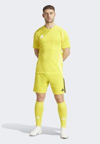 Geel sportief voetbalkostuum met een shirt met korte mouwen, bijpassende shorts, knielange sokken en witte voetbalschoenen met zwarte accenten.