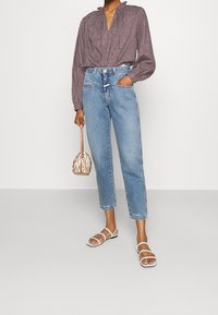 Blouse à motifs avec col flou, jean taille haute bleu clair, sandales blanches à lanières et sac en osier beige.