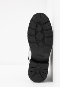 Suela de goma negra con un patrón de textura, mostrando la talla 37, vista inferior del zapato con acentos metálicos en la parte superior.